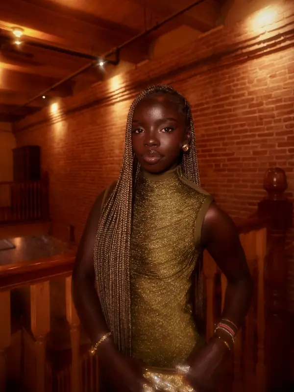 Joy in a gold knit dress with box braids against a brick wall